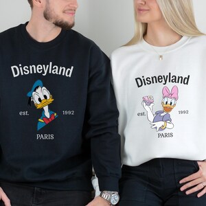 Pode incluir: Camisolas Disneyland azuis marinho e brancas. A camisola azul marinho apresenta o Pato Donald, e a camisola branca apresenta a Margarida. Ambas as camisolas t&ecirc;m o log&oacute;tipo da Disneyland, o texto "est. 1992 PARIS".