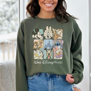 May include: Olive green sweatshirt featuring a grid of Disney characters including Mickey Mouse, Minnie Mouse, Donald Duck, and Stitch. The words "Walt Disney World" are printed below the grid in a white font. The sweatshirt is made of a soft material.
