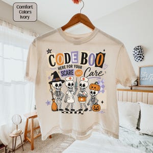 May include: An ivory t-shirt featuring a graphic with the text "CODE BOO HERE FOR YOUR SCARE AND CARE" and four skeletons dressed as a witch, a nurse, a medical professional with a syringe, and a pumpkin-headed figure with a jack-o'-lantern.