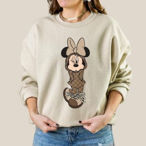 May include: Beige sweatshirt featuring a Minnie Mouse graphic. The cartoon character is dressed in a brown outfit with a bow and shoes. The outfit has a repeating pattern. The sweatshirt has long sleeves and a crew neck.