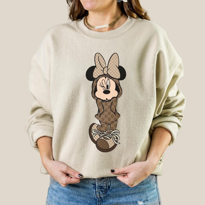 May include: Beige sweatshirt featuring a Minnie Mouse graphic. The cartoon character is dressed in a brown outfit with a bow and shoes. The outfit has a repeating pattern. The sweatshirt has long sleeves and a crew neck.