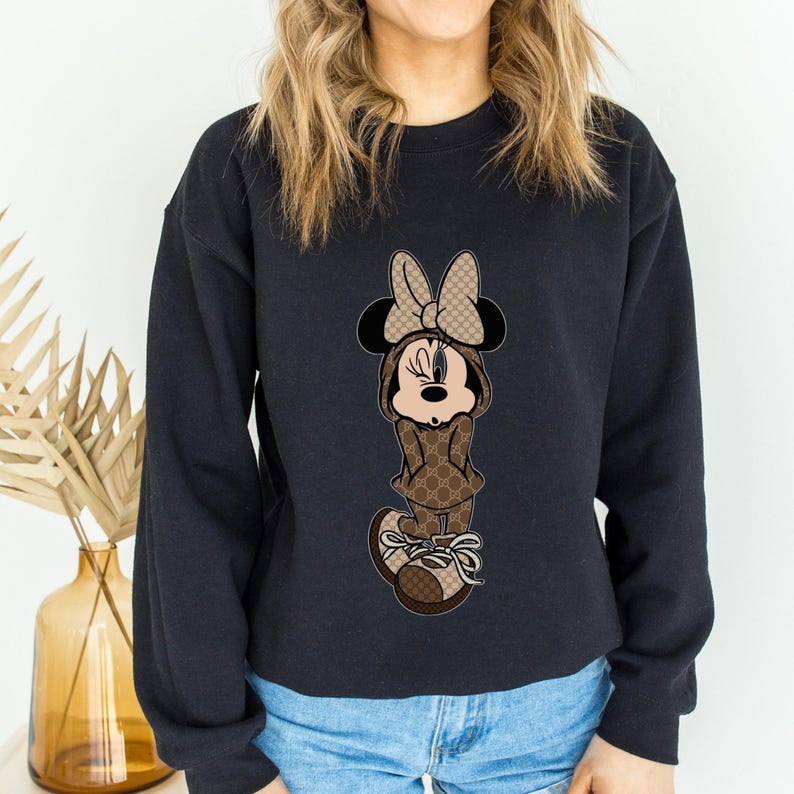 May include: Black crewneck sweatshirt featuring a cartoon Minnie Mouse character in a brown and tan patterned outfit. The character is wearing a matching bow and sneakers. The design is centered on the front of the sweatshirt.