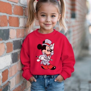 K&ouml;nnte beinhalten: Ein rotes Sweatshirt mit einem Cartoon-Motiv von Mickey Mouse in einem Zauberer-Outfit. Die Grafik zeigt Mickey Mouse in einem grauen Anzug, Zylinder und roter Fliege, der einen Gehstock h&auml;lt. Das Sweatshirt wird mit einer blauen Jeans kombiniert.