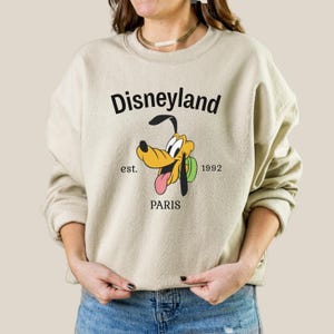 Pode incluir: Camisola bege com um gr&aacute;fico da Disneyland com Pluto, o c&atilde;o dos desenhos animados, e a palavra "PARIS" abaixo. A camisola tamb&eacute;m inclui o texto "est. 1992". O gr&aacute;fico &eacute; em preto, amarelo e rosa.