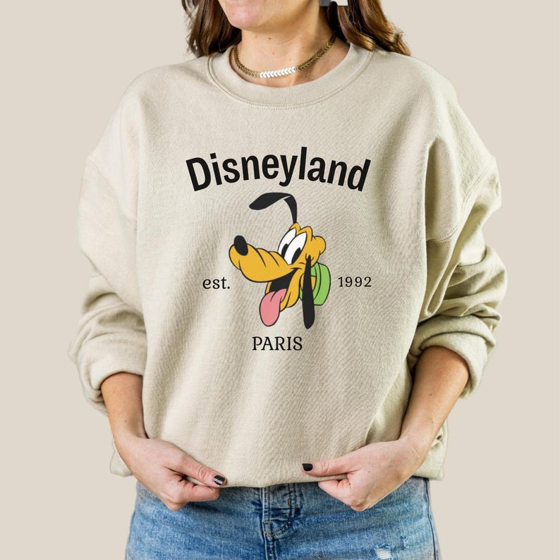 Pode incluir: Camisola bege com um gr&aacute;fico da Disneyland com Pluto, o c&atilde;o dos desenhos animados, e a palavra "PARIS" abaixo. A camisola tamb&eacute;m inclui o texto "est. 1992". O gr&aacute;fico &eacute; em preto, amarelo e rosa.