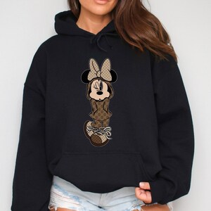 May include: Black hooded sweatshirt featuring a cartoon Minnie Mouse character. Minnie is depicted in a brown and tan patterned outfit, including a bow, dress, and sneakers. The design is centered on the front of the sweatshirt.