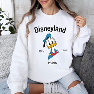 Pode incluir: Camisola branca de gola redonda com um desenho do Pato Donald usando um chap&eacute;u de marinheiro azul e gravata borboleta. A palavra "Disneyland" est&aacute; acima do desenho, com "est. 1992 PARIS" abaixo.