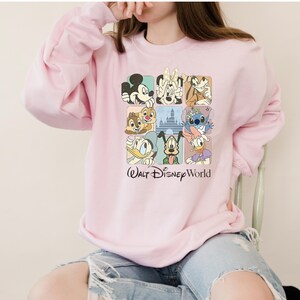 May include: A light pink sweatshirt featuring a grid of Disney characters including Mickey Mouse, Minnie Mouse, Goofy, Donald Duck, and Stitch. The words "Walt Disney World" are printed below the grid in black script.