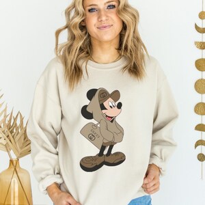 May include: Beige sweatshirt featuring a cartoon Mickey Mouse character in a brown hooded outfit and carrying a shopping bag with the Gucci logo. The sweatshirt is a neutral color, suitable for casual wear.