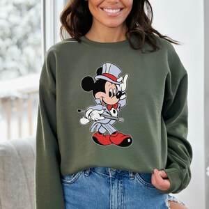 K&ouml;nnte beinhalten: Olivgr&uuml;nes Sweatshirt mit einem Cartoon-Motiv von Mickey Mouse in grauem Anzug, roter Schuhe und Zylinder. Mickey h&auml;lt einen Stock und winkt. Getragen von einer Person in blauen Jeans.