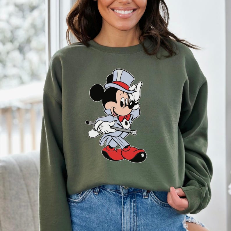 K&ouml;nnte beinhalten: Olivgr&uuml;nes Sweatshirt mit einem Cartoon-Motiv von Mickey Mouse in grauem Anzug, roter Schuhe und Zylinder. Mickey h&auml;lt einen Stock und winkt. Getragen von einer Person in blauen Jeans.