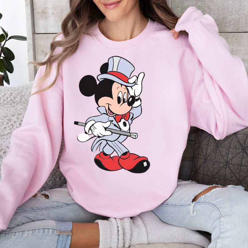 K&ouml;nnte beinhalten: Hellrosa Sweatshirt mit einem Cartoon-Bild von Mickey Mouse in einem Zauberer-Outfit. Mickey tr&auml;gt einen grauen Anzug, eine rote Fliege, rote Schuhe und einen Zylinder und h&auml;lt einen Zauberstab. Das Sweatshirt hat einen l&auml;ssigen Stil.