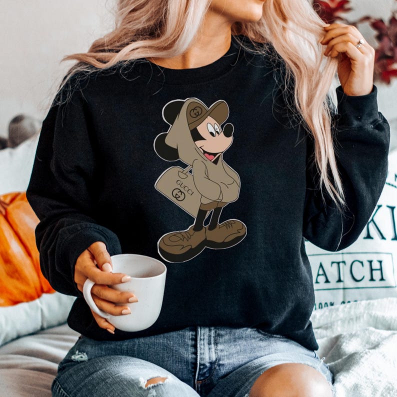 May include: Black sweatshirt featuring a cartoon Mickey Mouse character in a brown hoodie and hat, carrying a shopping bag with the Gucci logo. The character is wearing brown shoes. The sweatshirt is a solid black color.