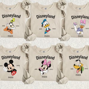 Pode incluir: Camisolas bege com personagens da Disney: Pateta, Pato Donald, Margarida, Mickey Mouse, Minnie Mouse e Pluto. Cada camisola tem a palavra "Disneyland" acima da personagem, com "est. 1992 PARIS" abaixo.