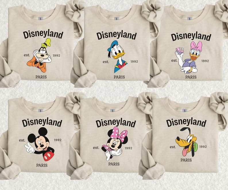 Pode incluir: Camisolas bege com personagens da Disney: Pateta, Pato Donald, Margarida, Mickey Mouse, Minnie Mouse e Pluto. Cada camisola tem a palavra "Disneyland" acima da personagem, com "est. 1992 PARIS" abaixo.