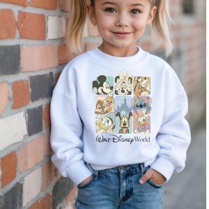 May include: White sweatshirt featuring a colorful graphic of Disney characters in square frames. The text "Walt Disney World" is printed below the graphic. The sweatshirt is worn by a young child.