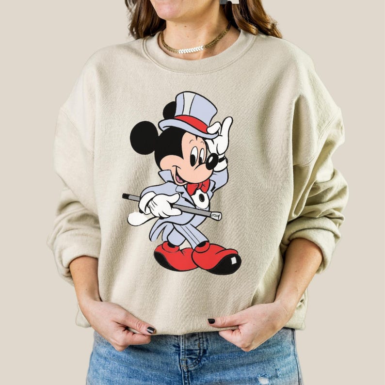 K&ouml;nnte beinhalten: Beiges Sweatshirt mit einer Cartoon-Illustration von Mickey Mouse in einem Zauberer-Outfit. Mickey ist in einem grauen Anzug, roter Fliege und roten Schuhen dargestellt, h&auml;lt einen Stock und tr&auml;gt einen Zylinder. Das Design befindet sich mittig auf dem Sweatshirt.