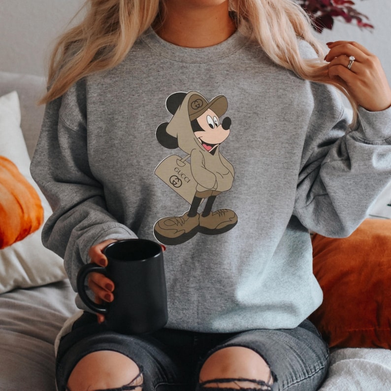 May include: A gray sweatshirt featuring a cartoon Mickey Mouse character in a brown hoodie and shoes, holding a shopping bag with the Gucci logo. The sweatshirt is a solid gray color.