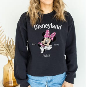 Pode incluir: Camisola preta de gola redonda com um desenho da Minnie Mouse com um la&ccedil;o rosa de bolinhas. A palavra "Disneyland" est&aacute; acima do desenho, com "est. 1992 PARIS" abaixo. Uma camisola de estilo cl&aacute;ssico.