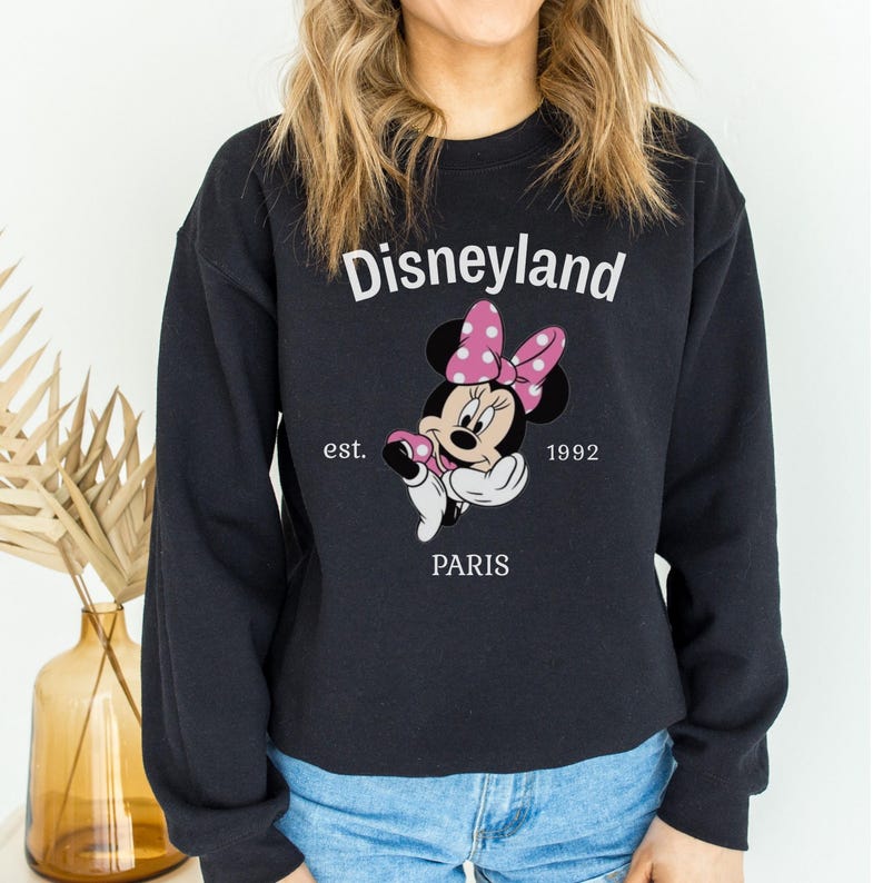 Pode incluir: Camisola preta de gola redonda com um desenho da Minnie Mouse com um la&ccedil;o rosa de bolinhas. A palavra "Disneyland" est&aacute; acima do desenho, com "est. 1992 PARIS" abaixo. Uma camisola de estilo cl&aacute;ssico.