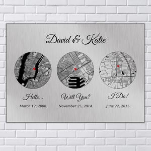 May include: A brushed metal plaque with three circular map illustrations, each with a red marker. The text "David & Katie" is above the maps. Below the maps are the phrases "Hello...", "Will You?", and "I Do!" with corresponding dates.