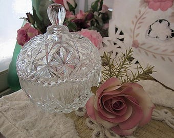 May include: A clear glass candy dish with a cut glass design and a lid. The dish is sitting on a white lace tablecloth with a pink rose.