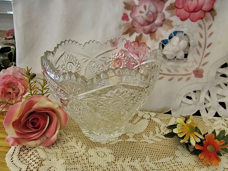 Peut inclure: Un bol en verre transparent avec un motif en verre taill&eacute;. Le bol est pos&eacute; sur une nappe en dentelle blanche avec des fleurs roses et jaunes.
