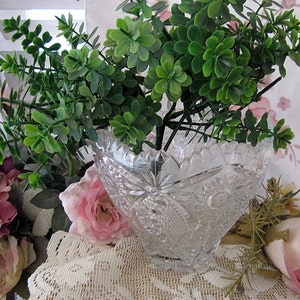 Peut inclure: Un bol en verre transparent avec un motif en verre taill&eacute;, rempli d'une plante verte feuillue. Le bol est pos&eacute; sur une nappe en dentelle blanche avec des fleurs roses.