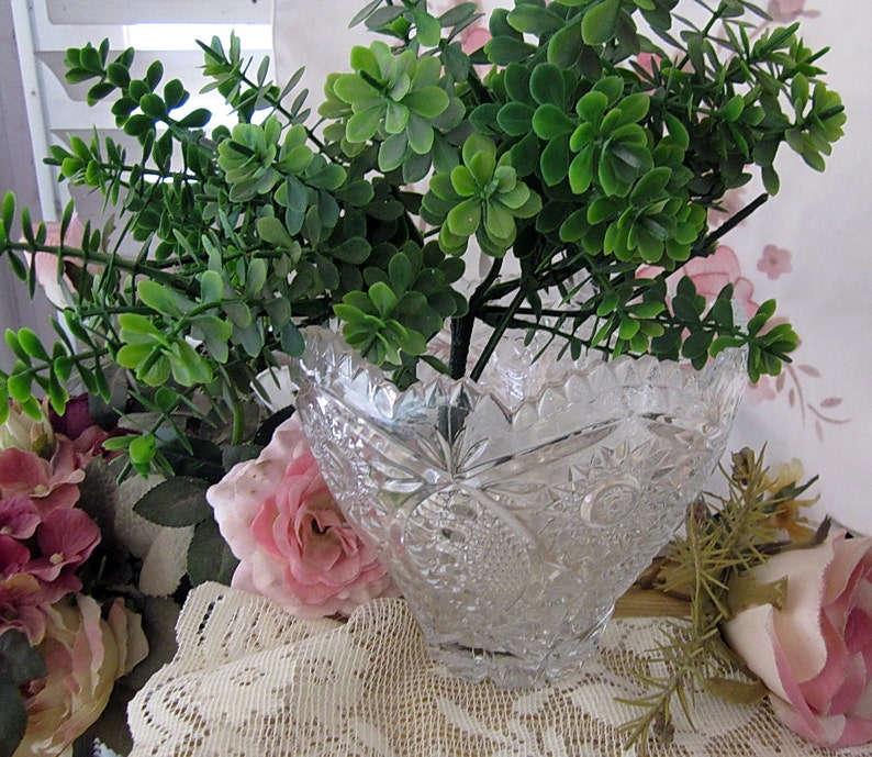 Peut inclure: Un bol en verre transparent avec un motif en verre taill&eacute;, rempli d'une plante verte feuillue. Le bol est pos&eacute; sur une nappe en dentelle blanche avec des fleurs roses.