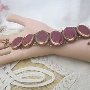 May include: A copper-toned bracelet featuring six oval, deep red gemstones. The stones are set in a copper frame and linked together with a delicate chain. The bracelet is secured with a toggle clasp.