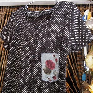 May include: A black and white polka dot shirt with a floral patch pocket. The shirt has a collar and short sleeves.