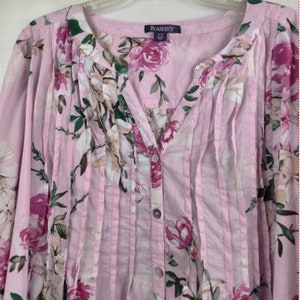 Könnte beinhalten: Eine rosa, geblümte Bluse mit Knöpfen, V-Ausschnitt und Faltendetail. Die Bluse hat einen Blumendruck mit rosa, weißen und grünen Blumen. Das Markenetikett lautet "ROMAN'S".