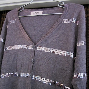 Può includere: Un cardigan grigio con paillettes argentate a strisce orizzontali. Il cardigan ha un collo a V e una chiusura con bottoni. L'etichetta recita "Miss Kioko"