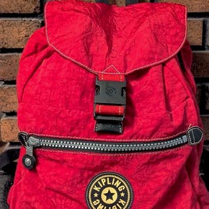 May include: A red backpack with a flap closure and a black buckle. The backpack has a zippered front pocket and a round logo patch with the brand name and a star. The bag has a black handle and straps.