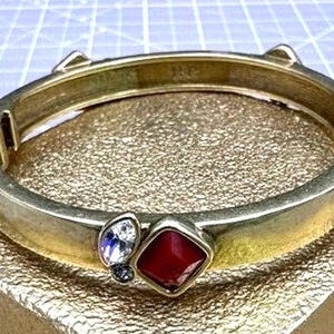May include: A gold bangle bracelet with red and clear stones. The bracelet is open and has a hinged clasp.