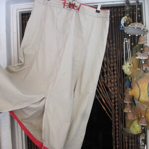 Puede incluir: Una falda larga de color blanco con una cremallera roja y un cordón rojo en la cintura. La falda está colgada de un gancho delante de una cortina de cuentas.