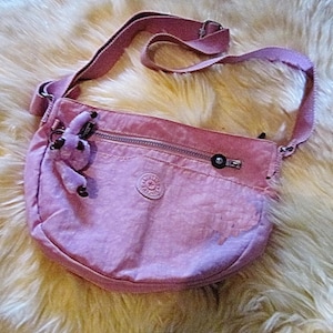 May include: A light pink crossbody bag with a matching adjustable strap. The bag features a zippered main compartment, a small front pocket, and a decorative charm. The bag is made of a durable, water-resistant material and has a round logo on the front.