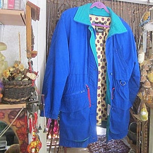 May include: A blue and teal jacket with a red zipper and a polka dot lining. The jacket has two pockets on the front and a drawstring waist.