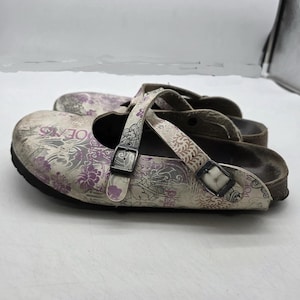 Może przedstawiać: Para chodaków z kwiatowym wzorem w odcieniach fioletu i bieli. Chodaki mają regulowane paski ze srebrnymi klamrami, a na boku widnieje napis "POEMS". Podeszwy są brązowe.