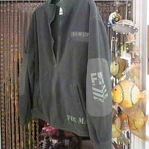 May include: A dark gray fleece zip-up jacket with a black zipper and a patch on the left sleeve that says "FM" in white letters. The jacket has a black zipper and a black collar. The text "LIMIT" is printed on the left chest pocket. The text "FOX MA" is printed on the bottom right side of the jacket.