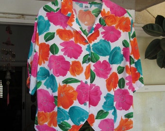 Könnte beinhalten: Weißes Hemd mit auffälligem Blumenmuster. Das Design umfasst große Blüten in Pink, Orange und Türkis, mit grünen Blättern. Das kurzärmlige Hemd hängt an einem schwarzen Kleiderbügel.