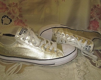 Puede incluir: Un par de zapatillas Converse bajas doradas metálicas con cordones blancos y puntera de goma blanca. Los zapatos tienen una raya negra a lo largo de la suela blanca y el logotipo de Converse All Star en la lengüeta y el lateral.