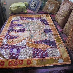 May include: A colourful patchwork tapestry featuring an elephant design with gold and silver sequins. The tapestry is displayed on a bed with pillows and a green frog plush toy.