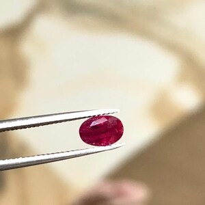 Pode incluir: Uma gema de rubi facetada em forma oval, segurada por uma pinça. O rubi é de cor vermelha profunda, com inclusões internas visíveis. A pinça é prateada e o fundo está desfocado.