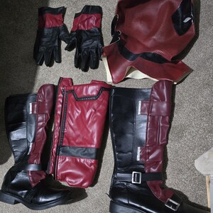Può includere: Un completo costume di Deadpool, con maschera, guanti, stivali e pettorina bordeaux e neri. Gli stivali hanno fibbie e cinturini. Il costume è appoggiato su una superficie neutra.