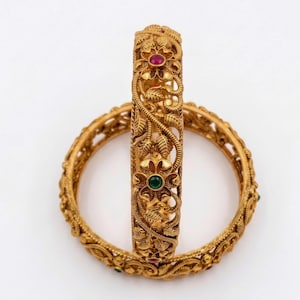 Peut inclure: Deux bracelets rigides dorés présentant un motif filigrane complexe de feuilles et de fleurs en relief. La surface métallique a une finition mate, et chaque centre floral est orné d'une petite pierre précieuse ronde incrustée, rouge ou verte.