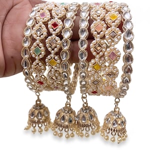 May include: A set of four gold-toned bangles featuring intricate metalwork, embedded with small white pearls and multi-colored square-cut stones. Each bangle is adorned with a dangling bell-shaped charm decorated with pearls and clear stones.
