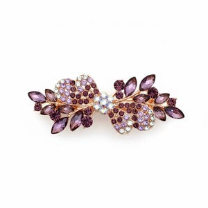 May include: A decorative hair barrette with a gold-tone metal base. The barrette features a floral design with purple and lavender-coloured rhinestones. The rhinestones are arranged in a leaf and flower pattern.