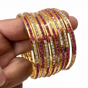 Peut inclure: Un ensemble de bracelets joncs dorés ornés de rangées alternées de cristaux blancs transparents et de pierres rose fuchsia éclatantes. Ces bracelets circulaires présentent une finition métallique polie et sont empilés.