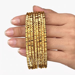 May include: A set of six gold bangles featuring a textured, faceted surface that reflects light. Each circular bracelet has a consistent width and a polished metallic finish, creating a repeating pattern of small, reflective geometric indentations.